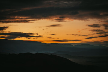 Scenic dawn mountain landscape with golden low clouds in valley among dark mountains silhouettes under sunset or sunrise sky. Vivid scenery with low clouds in mountain valley in illuminating color.