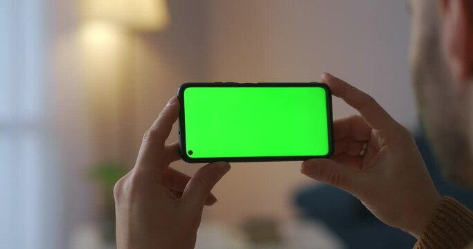man is holding smartphone horizontally and viewing video, closeup view, green screen for chroma key technology