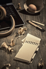 Creative writing, story board, writer's block. Concept still life on wood table. Crumpled paper. Open block note, surreal decor. Crumpled paper pages. Laptop, earphones, stone pyramid, chess figures.