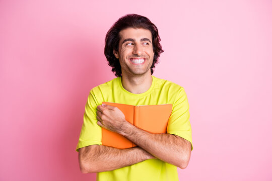 Photo of happy dreamy funky young man look empty space wear yellow t-shirt hug notebook isolated on pastel pink color background