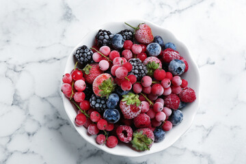 Mix of different frozen berries in bowl on white marble table, top view