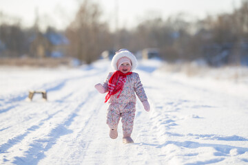 One happy beautiful baby girl in overalls and red scarf running on white snow covered country road...