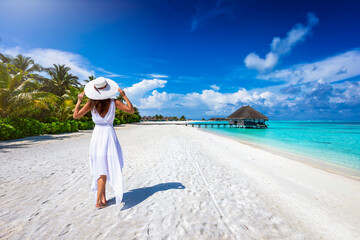 A woman with a white dress on holiday in the Maldives walks down a beach with palm trees and...