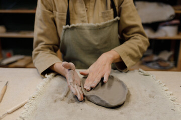 A young Caucasian woman makes sides with her hands for a plate of clay in a pottery workshop. Beautiful dishes with their own hands. Manual labor. Creative masterclass