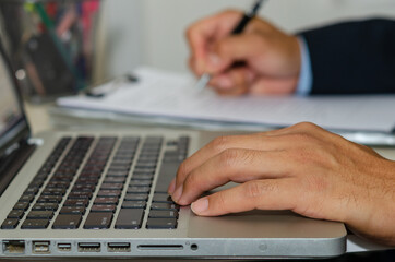 business man hand typing a computer and holding a pen to write business documents on the desk