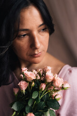 young thirtysomething woman portrait with pink roses and natural light vertical close up. concept of midlife sophisticated woman. confident femininity. serious expression and soft light. pink dress
