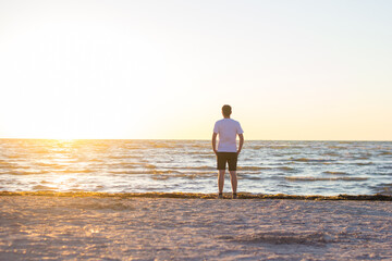 Fototapeta na wymiar Rear view of a young man in a white shirt and brown shorts standing on empty beach at sunrise. A male stands on the shores of a calm morning sea and looking at sunrise.