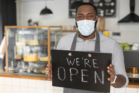 A cheerful young African American male waiter wearing mask holding in hands open sign indoors in own bakery shop. Reopening bakehouse after lockdown, pandemic, epidemic