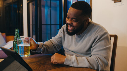 Middle-aged African American male drinking beer having fun happy night party event online celebration via video call in living room at home. Social distancing, quarantine for coronavirus prevention.
