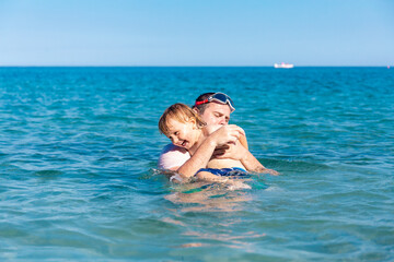 Fototapeta na wymiar Father and little son having fun swimming and playing together in sea water at summer holidays. Family bonding, togetherness. Seaside vacation.