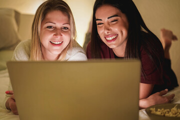 Two young girl friends using laptop computer together on bed during coronavirus lockdown isolation - Focus on left girl