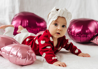 Stylish infant in trendy white headband on bed with balloons. .Cute baby girl in red sweater with...