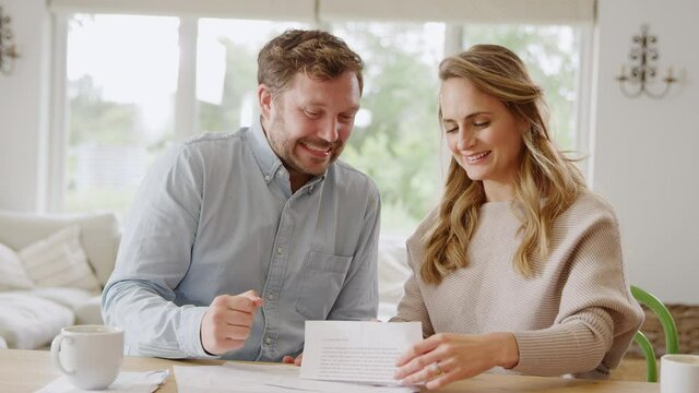 Excited couple sitting at table at home opening letter containing good financial news - shot in slow motion
