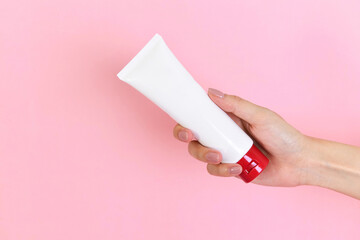 Close-up of young female's right hand holding blank white squeeze flexible plastic tube with a red lid isolated on a pink background. Cream, lotion, gel, facial foam, skincare, cosmetic packaging