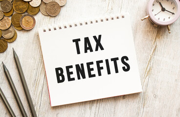 TAX BENEFITS text on a sheet of notepad. Coins are scattered, pencils on a gray wooden background.