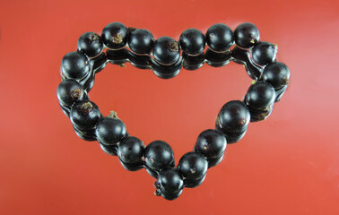 Valentine card with heart Black currant berries  on  red mirror wet surface, reflected in water.  Symbol of Valentine's day, romantic holiday, celebration of love and affection.