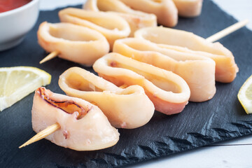 Fried squid rings on a skewer with tomato sauce and lemon. Black slate stand, close up.