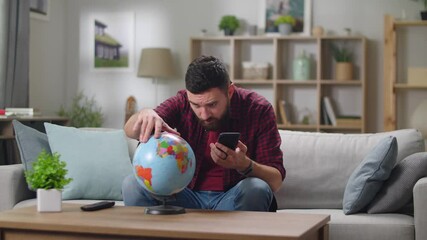 Young handsome guy looking for a country on the globe and in the smartphone while sitting at home in the living room - Powered by Adobe
