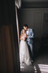 Fototapeta premium Wedding portrait of stylish groom in gray plaid suit and cute blonde bride with curly hair near the window with curtains in the hotel room. Interior photography.