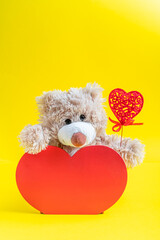 Lovely brown teddy bear toy sitting in wooden red heart shaped decor with carved heart decoration in its paw as a gift on saint valentine's day or other special date. Vertical orientation image