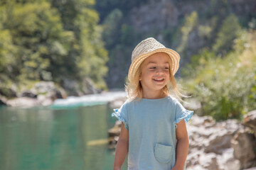 Portrait of little adorable blonde girl, who sitting on the river coast on stones. Child enjoy view of Tara river canyon, mountains landscape.