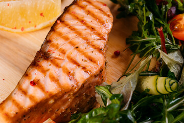 Grilled salmon steak on a wooden board with a fresh healthy salad, lemon, vegetables, herbs and spices on a dark concrete background. 