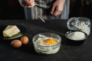 The process of preparing the dough. Ingredients for cookies on a dark background. In the women's hands, a spoon of salt is poured into a bowl with egg-flour mixture.
