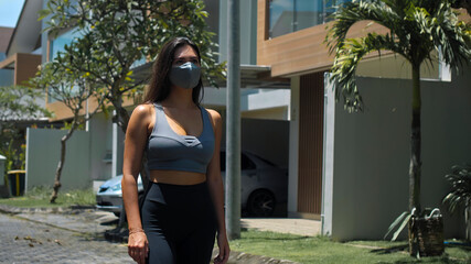 European brunette sports girl walks along an empty city street with green trees on the background alone in a gray protective mask