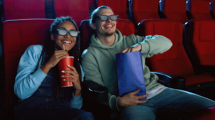 Happy young couple wearing 3d glasses smiling, while watching a movie together in cinema auditorium, eating popcorn and drinking soda