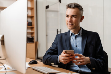 Pleased grey man using mobile phone while working with computer