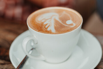 Fototapeta premium Closeup shot of white cup filled with aromatic coffee with heart shape latte art foam on wood table near window with light shade on tabletop at cafe with spoon besides it 
