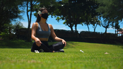 Young European brunette girl practices alone yoga in nature sitting on green grass in lotus pose wearing a black protective mask