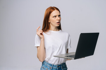 Thinking young woman student using laptop computer and looking away on isolated gray background. Pretty redhead lady model emotionally showing facial expressions in studio, copy space.