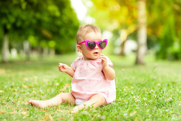 baby girl sitting on the green grass in a pink bodysuit and bright glasses, walking in the fresh air