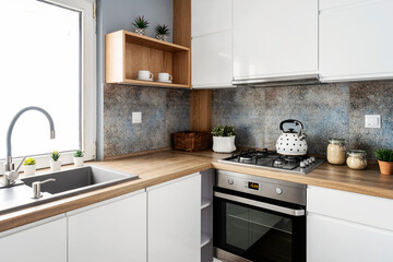 Small white kitchen with stylish tiles, wooden cabinets and sink with faucet near the window. Scandinavian interior of room in apartment.