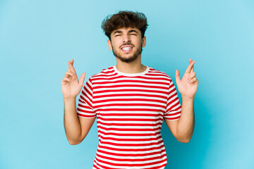 Young arab man on blue background crossing fingers for having luck