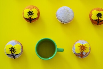 Rows of muffins decorated with powdered sugar and yellow flower shaped icing on a yellow background next to a cup of tea