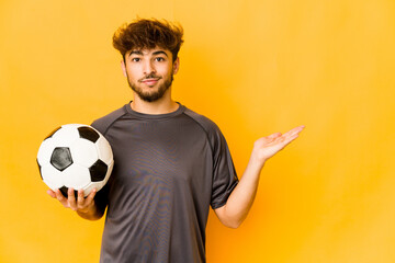Young soccer player indian man showing a copy space on a palm and holding another hand on waist.