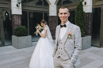 Naklejka premium Stylish groom in a gray suit and black bow-tie and the bride in a white dress with a bouquet in their hands are out of focus on the background of buildings in the city. Wedding portrait.