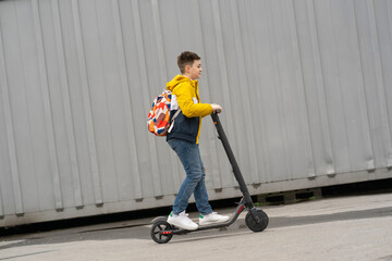 Fototapeta na wymiar Modern teenager with backpack rides on electric scooter. Boy comes back from school