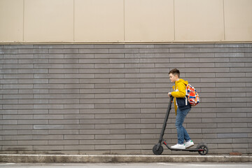 Modern teenager with backpack rides on electric scooter on brick wall background. Boy comes back from school