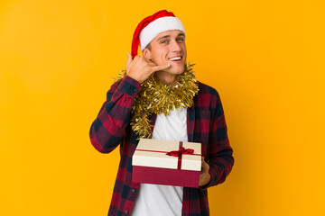 Young caucasian man with christmas hat holding a present isolated on yellow background