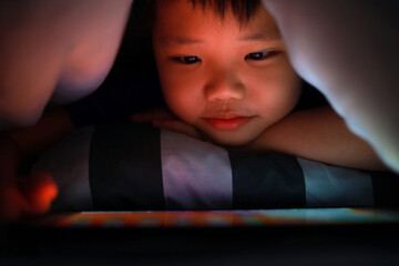 Asian boy hiding in the blanket on bed and playing digital tablet. Soft focus image.
