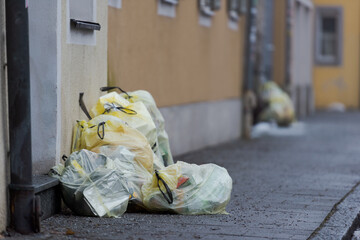 Yellow plastic sacks containing plastic garbage piled up near front-doors on sidewalk of street for waste collection in old town with stone houses