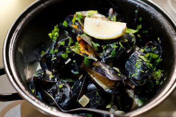 High angle view of steamed mussels in a metal bowl with sliced lemon and parsley twig. Selective focus on mussels.