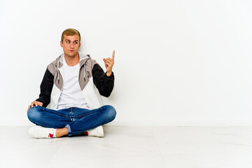 Young caucasian man sitting on the floor pointing with finger at you as if inviting come closer.
