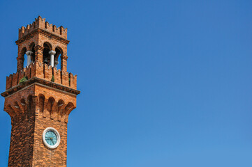 Detail of clock tower made of bricks with sunny blue sky in the background at Murano, a small and...