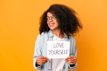 Young african american curly woman holding a love yourself placard looks aside smiling, cheerful and pleasant.