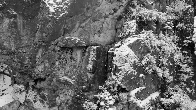 Black and white aerial of snow covered rock in the winter forest while snowflakes falling out of the sky
