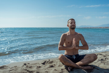 Fototapeta na wymiar Meditation on beautiful sandy beach. Handsome young skinny man meditating and doing yoga on beach. 